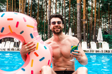 Happy young man holding inflatable ring and cocktail while resting in the swimming pool outdoors....