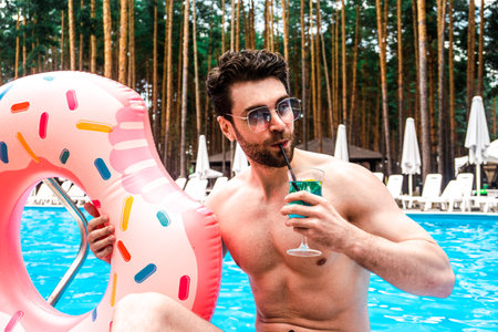 Young man in sunglasses drinking cocktail while enjoying rest near the pool, holding inflatable ring outdoor. Summertime, holidays, lifestyle conceptの写真素材