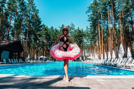 Happy pretty lady in black swimsuit holding inflatable ring near swimming pool outdoors. Lady in hat...