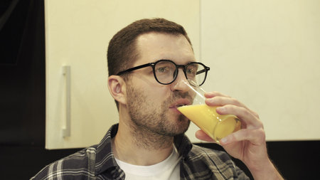 Calm young man wearing glasses and drinking orange juice, standing at kitchen