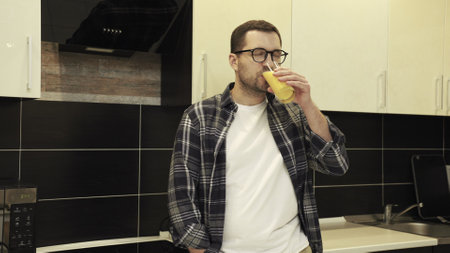 Young man in glasses standing at the kitchen while drinking fresh orange juiceの写真素材