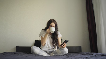 Young woman in pajamas drinking coffee while typing on the phone at homeの写真素材