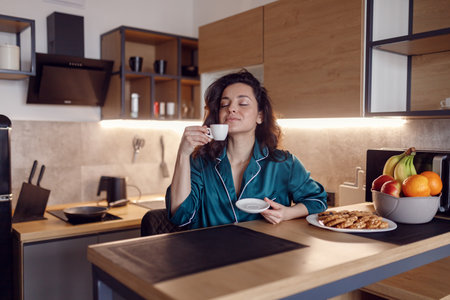 Beautiful young woman in silk robe enjoying the aroma of coffee with closed eyes while sitting resting at the kitchen at home. Morning routine, domestic lifeの写真素材