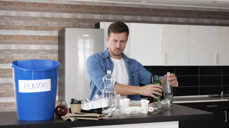 Young man in kitchen during quarantine. Sorting plastic bottle separated from other trash and put it into blue bucket. Responsible using. Take care of environment. Zero waste process.の写真素材