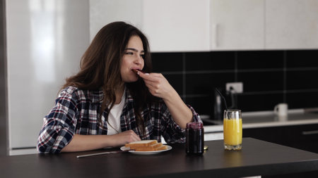 Young woman in kitchen during quarantine. Girl sit at table and eat toast bread with jam. Enjoying breakfast or morning meal with drinking orange juice.の写真素材