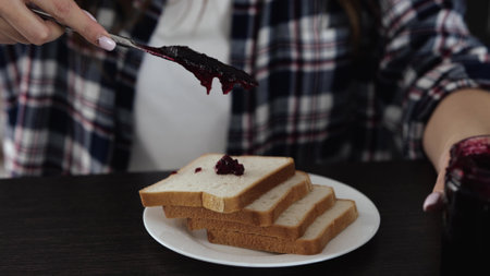 Young woman in kitchen during quarantine. Close up motion of woman using fork for putting some red jam on bread. Sweet toast ready to be eaten.の写真素材