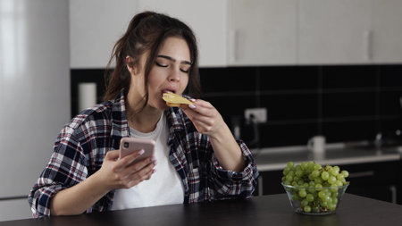 Woman in kitchen during quarantine. Sit at table and eat sandwich with cheese. Chatting on smartphone. Social media online. Breakfast time.の写真素材