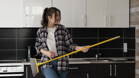 Young woman in kitchen during quarantine. Stand alone and pretend to play on mop as on guitar. Have fun, dancing and singing out loud during cleaning. Slow motion.の写真素材