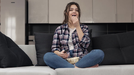 Young woman in kitchen during quarantine. Sit on sofa and watch tv or movie. Switching tv channels and eating popcorn. Laughing and have fun.の写真素材