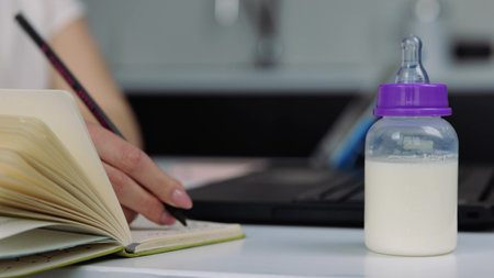 Young woman in kitchen during quarantine. Female hands typing on laptops keyboard and writing notes in notebook. Blurred background. Take baby milk bottle and walk away.の写真素材