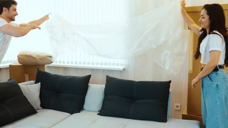 Young couple move into new apartment. Slow motion of taking cover out from sofa and walking to it. Sit down together and enjoy their time in new apartment.の写真素材