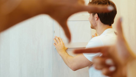 Young couple move into new apartment. Guy put on frame on wall and look back on his girlfriend with smile. Blureed womans hands in rectangle shape upon frame.の写真素材