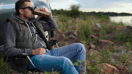 A man is resting on a car trip on the coast of a lake, ocean or sea. He has a seat in a folding chair. He is holding a thermos or thermo mugの写真素材
