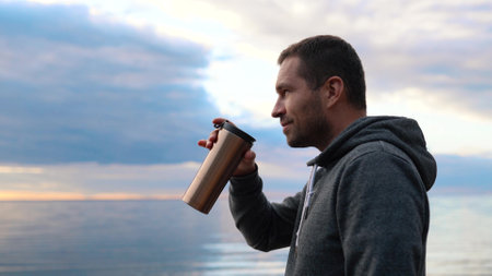A man drinking coffee or tea from a warm thermos against the backdrop of a sunset in a calm sea or oceanの写真素材