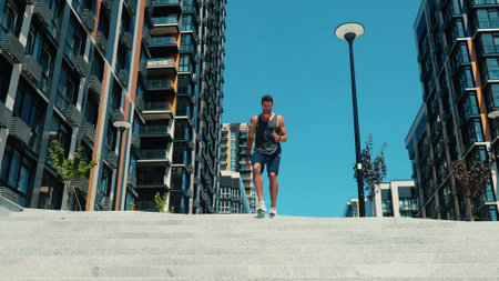 Young man exercising outside. Slow motion of guy starts jogging down steps listening to music. Running to video camera and jumping over it. Beautiful sunny day with no clouds. Blue sky.の写真素材