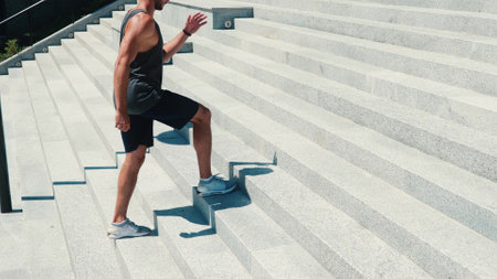 Young man exercising outside. Active strong guy doing squat moving skipping one step. Listen to music in white earphones. Enjoy outside training alone. Sunny beautiful day.の写真素材