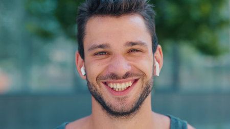Young man exercising outside. Camera moves back from handsome positive cheerful guy smiling out loud. Listen to music through white earphones.の写真素材