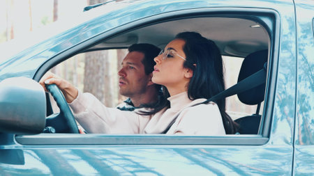 Young man and woman riding together in car in forest. Instructor helps female student to drive right way. Girl holding hands on steering wheel and driving.の写真素材