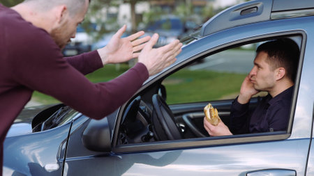 Young man inside car. Businessman eating burger and talking on phone. Another guy stands beside car and scream with argue on man inside. Businessman shows sign to him.の写真素材