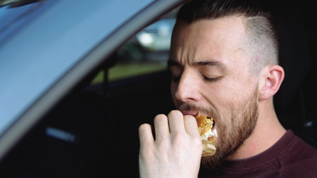 Young man insdie car. Portrait of hipster guy chewing tasty delicious hamburger and drink from paper cup. Enjoy take away breakfast or lunch in car.の写真素材