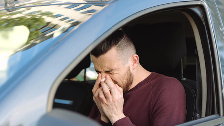 Young man inside car. Guy suffers from sneezing and terrible headache. Sits alone with symptoms. Sneezing into white tissue.の写真素材