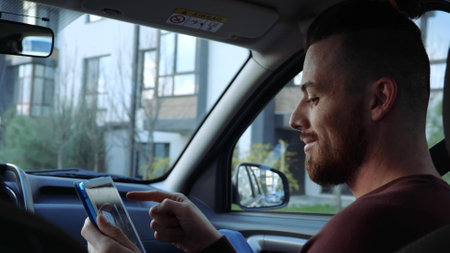 Young man inside car. Sitting on front seat and playing on tablet. Touching screen and having fun during ride in taxi cab or private car.の写真素材