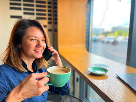 Close up of happy beautiful woman talking on cellphone while sitting at window in restaurant looking away and drinking coffee. Leisure concept. Female having cellphone call.の写真素材