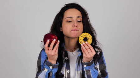 Young woman holding apple and cookie, choice difficulty. Isolated on grey background. Healthy, junk food concept.の写真素材