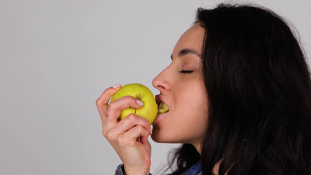 Smiling lady enjoying fresh yellow apple while biting it, isolated on grey background, copy space,...