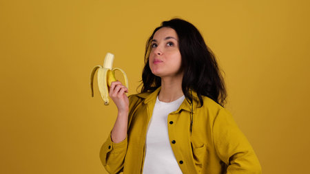 Smiling beautiful woman enjoying while eating banana, posing in studio, isolated on yellow...