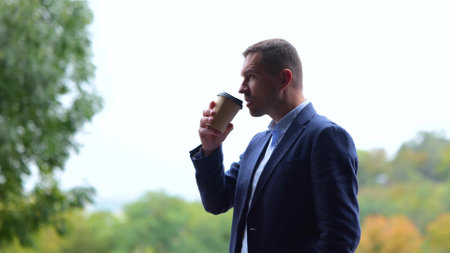 Portrait of one successful male professional standing outdoors in nature drinking coffee from paper cup. Caucasian man in suit with cup of coffee-to-go in park. Side viewの写真素材