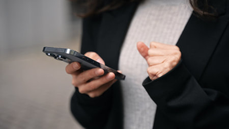 Caucasian female standing outdoors around grey modern buildings, working on her phone, smiling, touching her long dark hair. Close up.の写真素材
