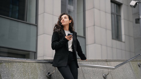 Young Caucasian businesswoman wearing office-style jacket standing next to office buildings,...