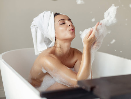 Caucasian cheerful woman sitting in bathtub with towel on head playing with soap blowing on foam...