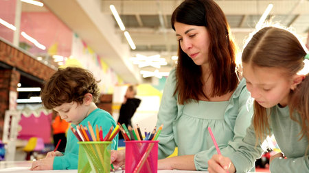 Young mother with children spending time in the game zone while painting with pencils together. Slow...