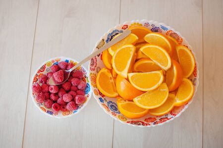 Big bowl with sliced oranges and small with raspberries and spoon on a light wooden background.の写真素材