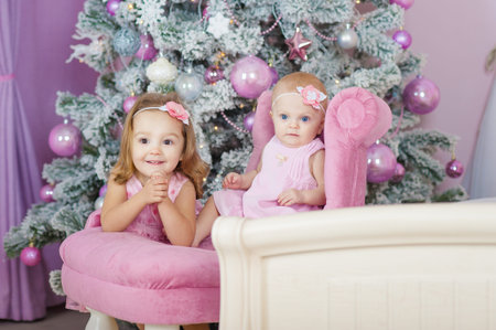 Little girls in beautiful dresses, sitting on a chair in the beautiful Christmas decorations. Two little sisters. New Year preparation. Happy girl and family.の写真素材