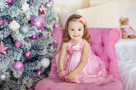 Very nice charming little girl blonde in pink dress sitting on a child's armchair and laughs loudly on the background of Christmas trees in bright interior of the house.の写真素材