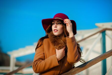Beautiful fashionable woman in a hat and coat posing against the blue skyの写真素材