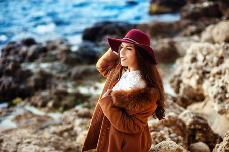 Cheerful lovely young woman in maroon felt hat and drape coat standing and laughing on pier in springの写真素材