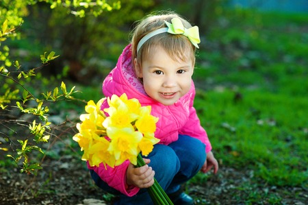Girl With a bow on his hair sitting on the grass and holding a bouquet of yellow daffodils. A child walks outside in warm weather.の写真素材