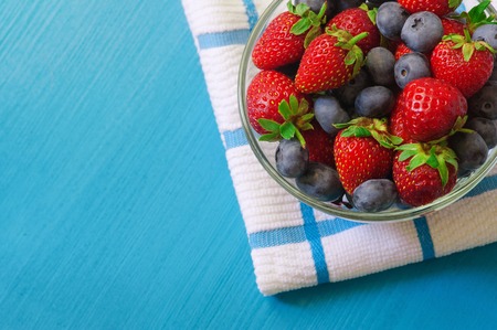 Glass bowl with assortment berries blueberries, strawberries and blackberries at white textile napkin over blue wooden table. Natural day light. Top view.の写真素材
