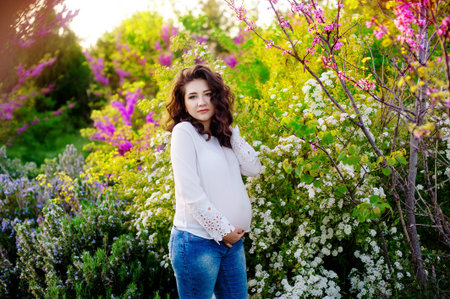 Beautiful young happy pregnant woman staying in fashion clothes in flower park touching her belly...