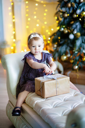 cute little girl in lilac dress sitting in a chair and opens box with present for background Christmas treeの写真素材