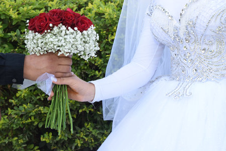 the hands of the newlyweds, the bride and groom in white at the wedding with a bouquet of scarlet...
