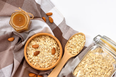 Oatmeal in a wooden bowl with almonds and pieces of banana ready to cook, honey, scattered oat flakes and spoon on white background. View from above.の写真素材
