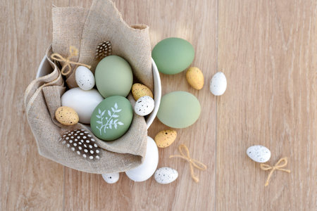 Stylish decorated easter eggs in a white bowl on linen cloth on a light wooden table. Top view. With...