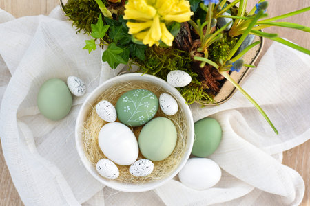 Stylish decorated easter eggs in a white bowl and spring primroses in a pot on linen cloth. Top...