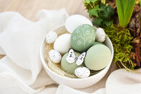 Stylish decorated easter eggs in a white bowl on linen cloth on a light wooden table. Happy Easter conceptの写真素材