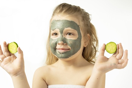 Funny little girl with a cosmetic mask of clay in a bath towel holding a piece of cucumber, like a mask, the concept of beauty and health, portrait indoors closeup.の写真素材